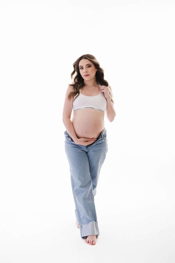 Pregnant woman in a white Calvin Klein bra and blue jeans stands barefoot on a white background, holding her belly with one hand and her bra strap with the other.