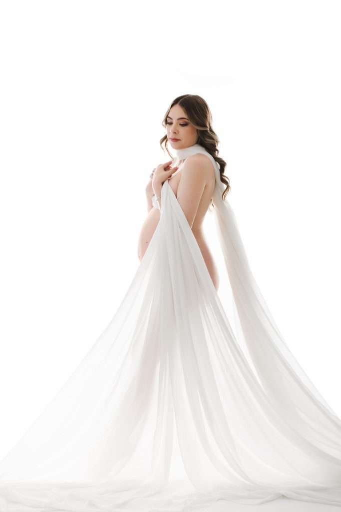 Pregnant woman standing in profile, draped in white translucent fabric, against a white background.