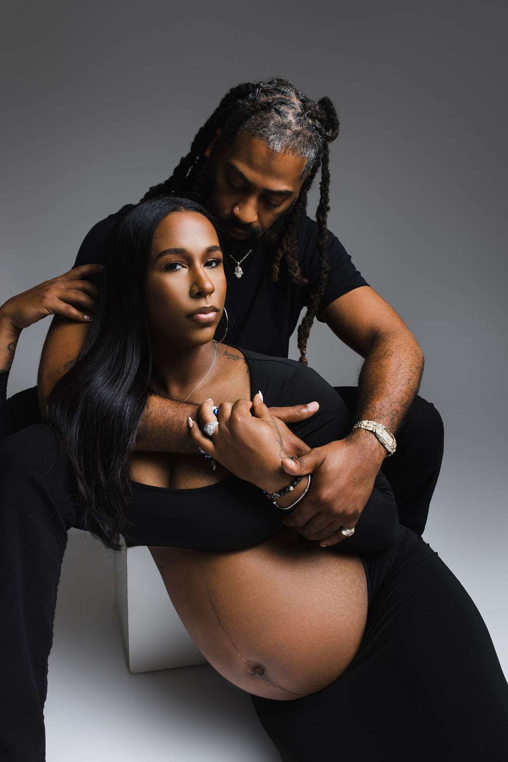 A pregnant woman sits with her bare belly exposed while a man embraces her from behind; both wear black clothing and have serious expressions.