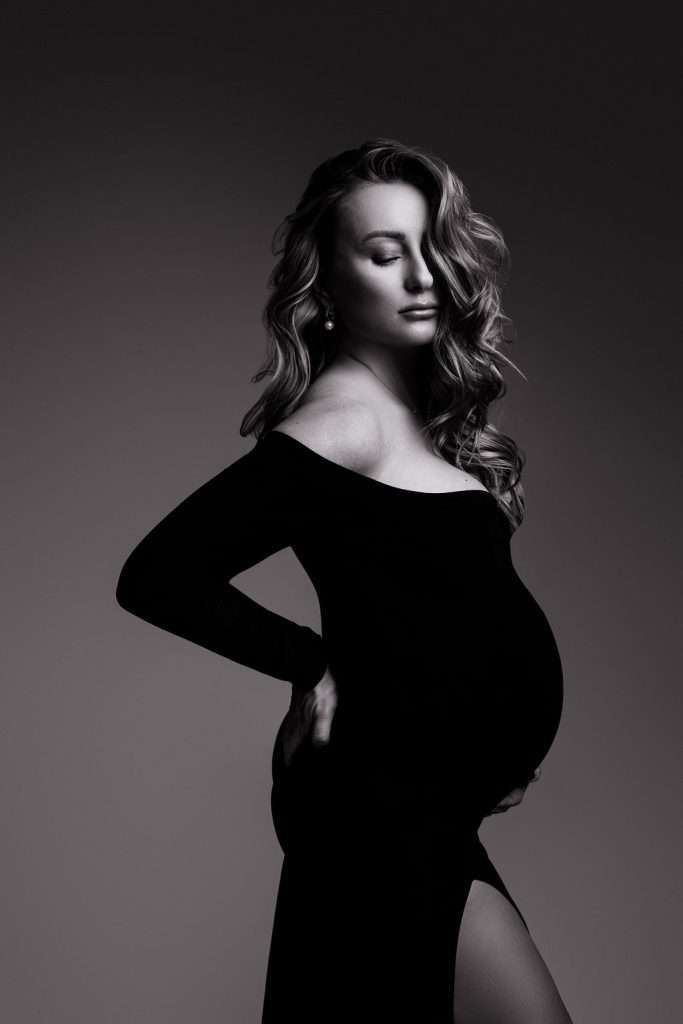 A pregnant woman in a long-sleeved, off-the-shoulder black dress poses with eyes closed against a plain background.