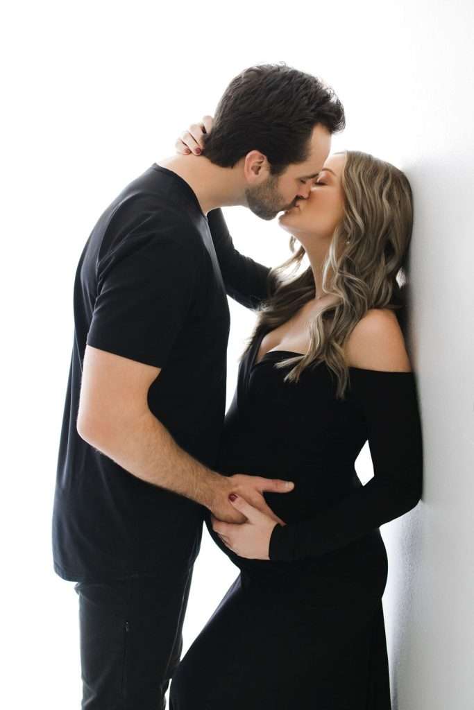 A man and a pregnant woman wearing black outfits kiss while standing against a white background. The man touches the woman's baby bump.