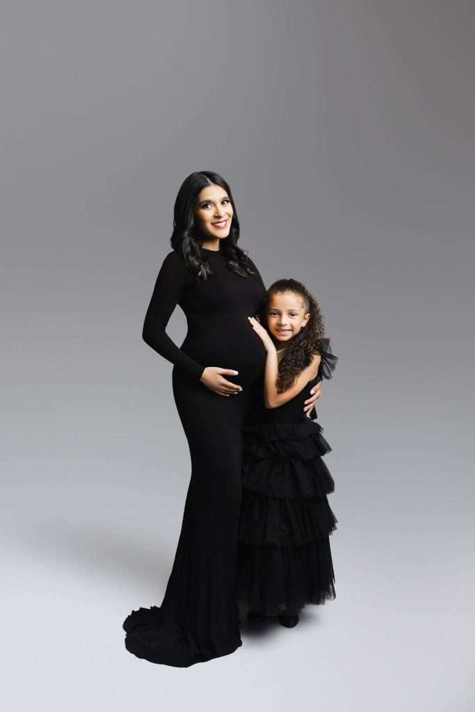 A pregnant woman in a long black dress stands with a young girl in a matching black dress, both smiling at the camera against a plain gray background.