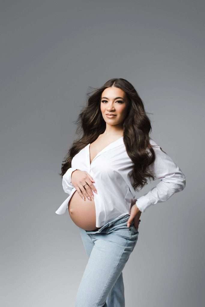 Pregnant woman with long dark hair poses in an open white shirt and light blue jeans against a plain gray background.