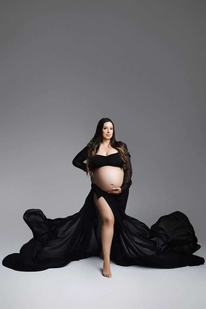 Pregnant woman in a black flowing gown poses barefoot against a plain gray background, with one hand on her hip and the other resting on her belly.