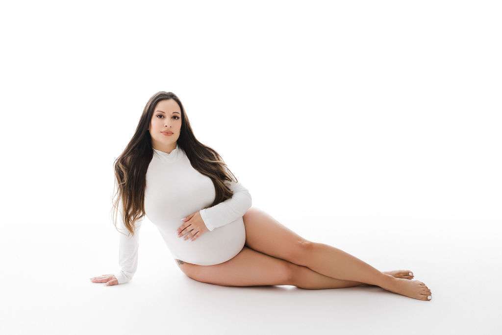 Pregnant woman in a white long-sleeve top sits on the floor with one hand on her belly, against a white background.