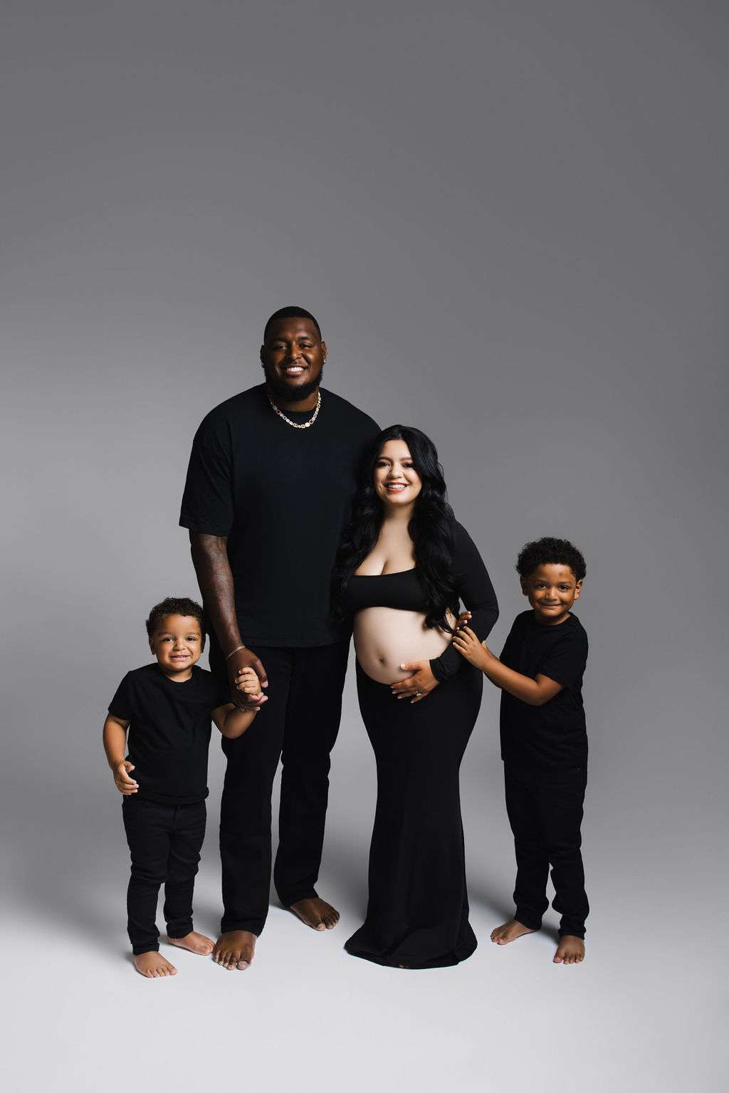 A family of four stands together, with the mother showing her pregnant belly. All are barefoot and wearing black clothes, posing against a plain gray background.