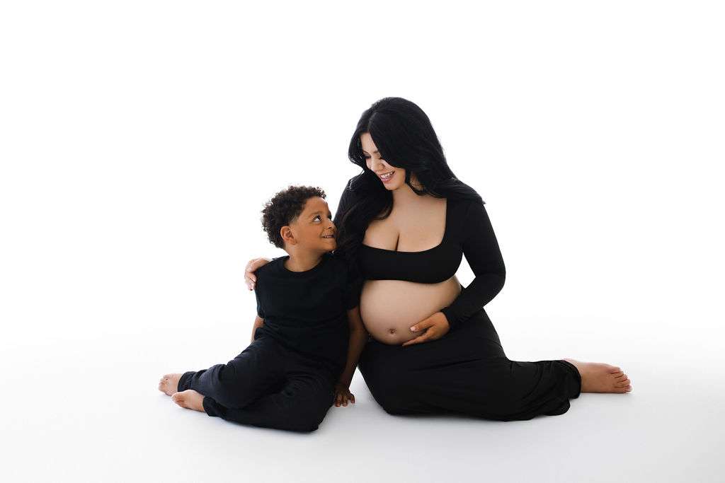 A pregnant woman in a black dress sits on the floor, smiling and holding her belly, with a young boy in black sitting beside her.