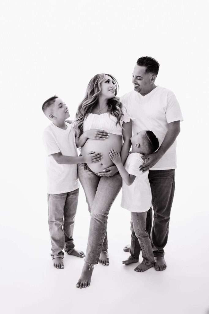 A pregnant woman stands with her partner and two young boys, all touching her belly and smiling, dressed in white shirts and jeans against a plain background.