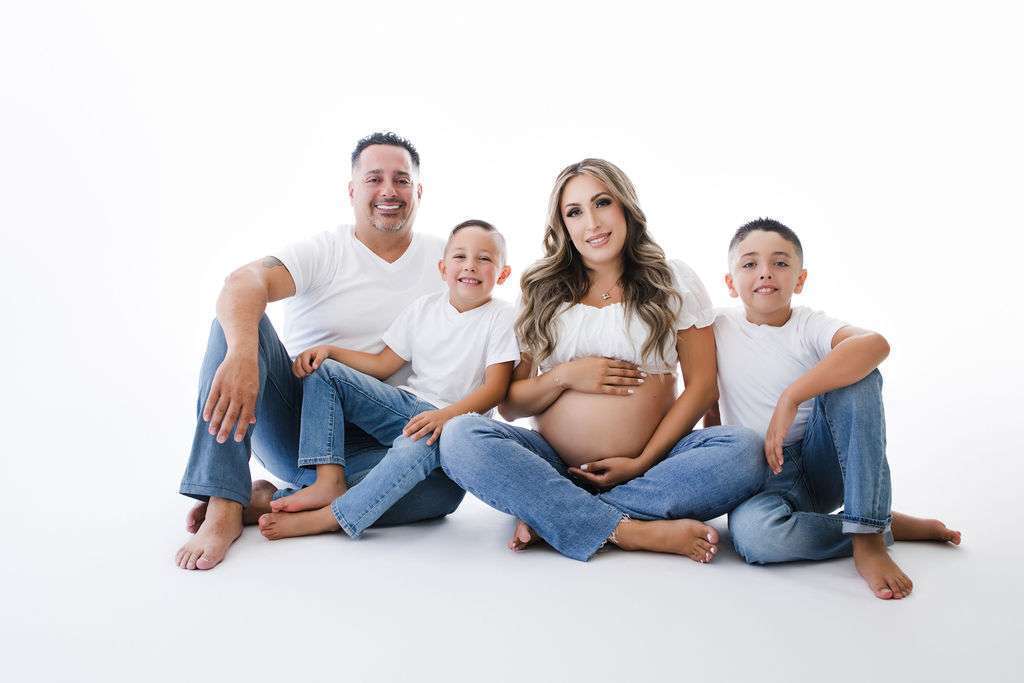A family of four sits barefoot on the floor in white tops and jeans. The mother, who is pregnant, sits in the center with her partner and two sons beside her, all smiling at the camera.