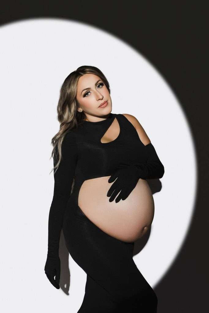 A pregnant woman in a black dress and gloves poses against a round spotlight, with her hand resting on her bare belly.