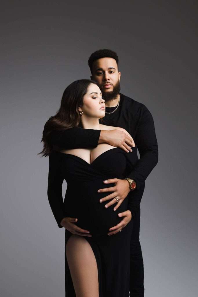 A couple poses against a gray background; the woman, visibly pregnant, wears an off-shoulder black dress while the man stands behind her, embracing her with his hands on her belly.