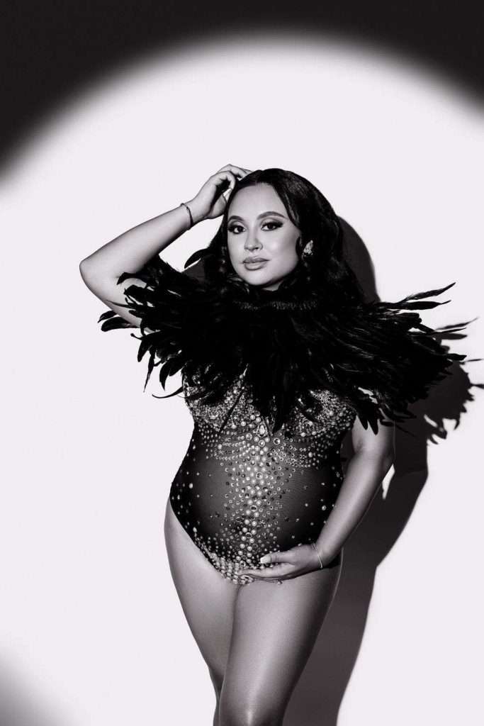 A person poses in a beaded bodysuit with a feather collar, holding their pregnant belly, standing under a spotlight against a plain background.