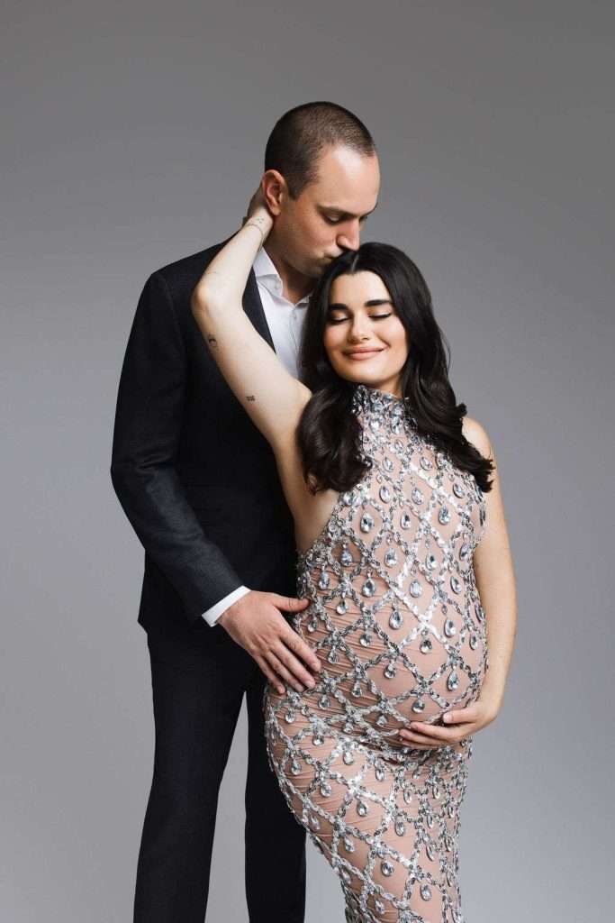 A man in a suit stands behind a pregnant woman in an embellished dress, gently kissing her head while she holds her belly and smiles with her eyes closed.