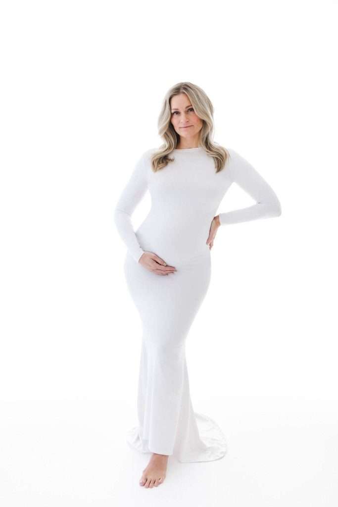A pregnant woman in a long-sleeved, white dress stands barefoot on a white background, one hand on her hip and the other on her belly.