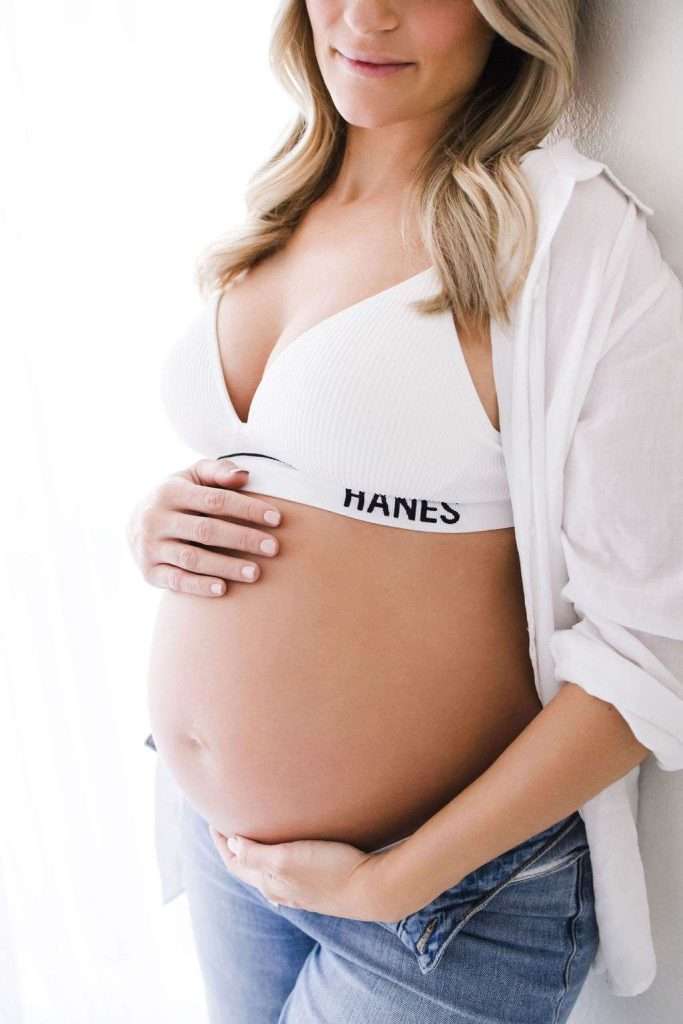 Pregnant woman in a white bra and open shirt gently cradling her bare belly, standing by a bright window.
