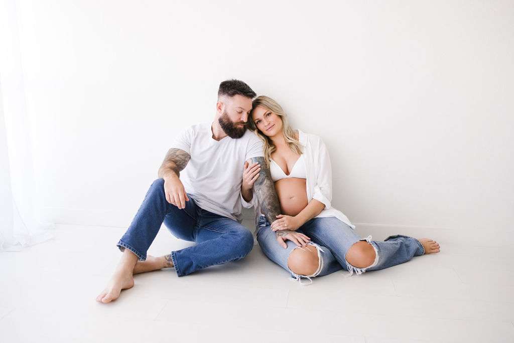 A man and a pregnant woman sit on the floor against a white wall. Both wear white tops and ripped jeans. The woman rests her head on the man's shoulder.
