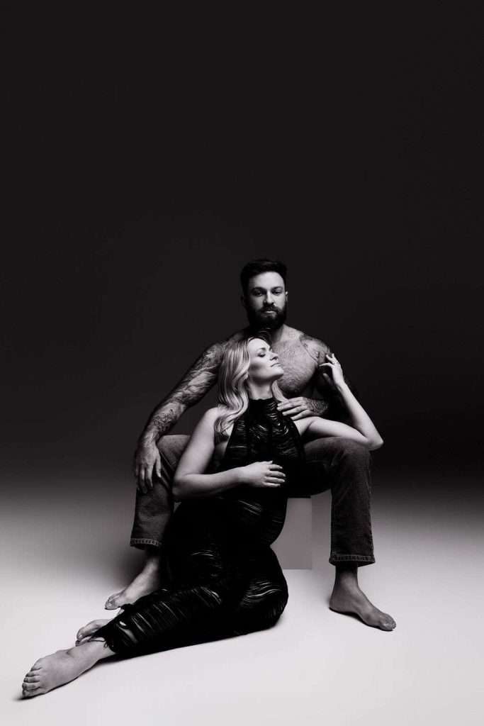 A pregnant woman in a black dress sits on the floor, leaning against a seated, shirtless man with tattoos. Both have neutral expressions, and the background fades from dark to light.
