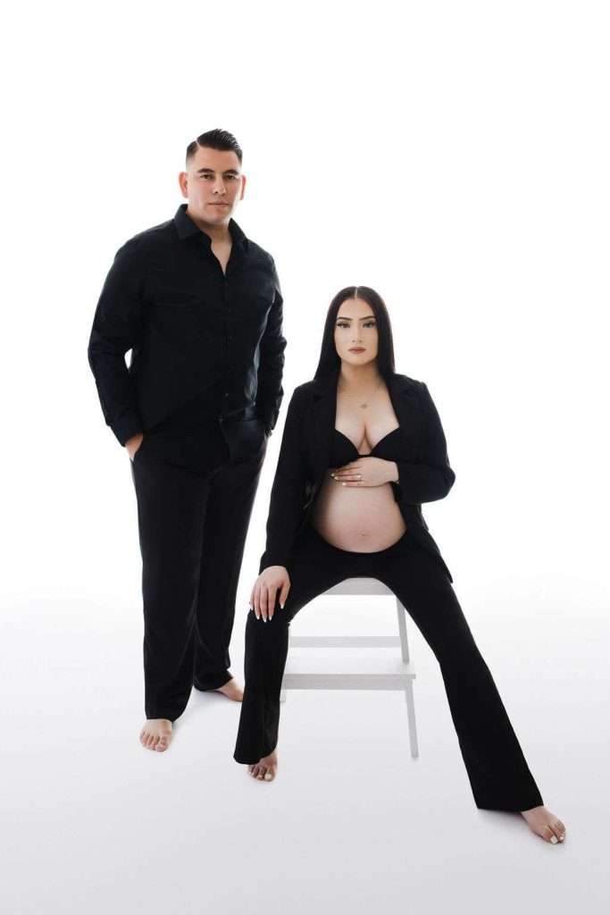 A barefoot couple in black outfits poses in a studio; the pregnant woman sits on a stool with one hand on her belly, while the man stands with one hand in his pocket.