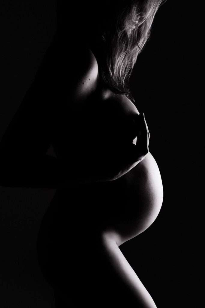 Silhouette of a pregnant woman in profile, one hand resting on her chest and the other supporting her lower back, with dramatic lighting highlighting her belly.