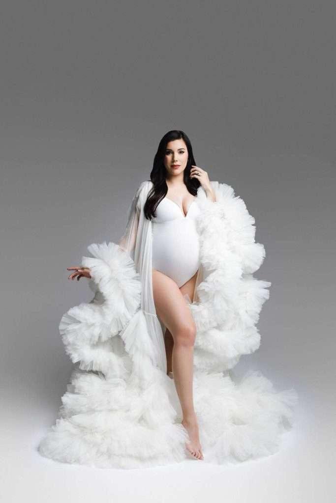 A pregnant woman in a white bodysuit and a long, ruffled sheer robe poses confidently against a gradient gray background.
