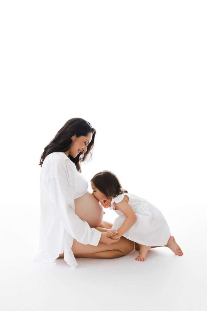 A pregnant woman sits on the floor, smiling, while a young girl in a white dress kisses her bare belly against a white background.