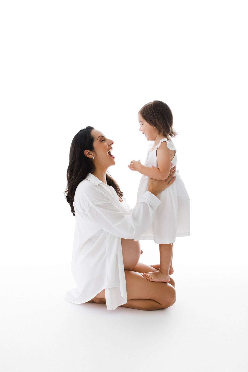 A woman kneels on the floor, smiling at a young girl in a white dress standing in front of her against a white background.