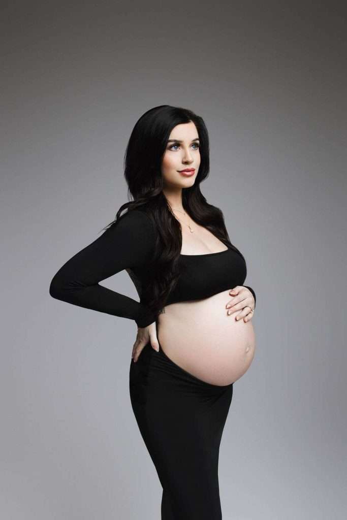 Pregnant woman with long dark hair in a black outfit stands in profile, one hand on her back and the other on her bare belly, against a plain gray background.
