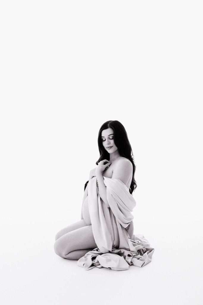 A pregnant woman kneels on the floor, draped in a light cloth, with a serene expression and a white background.