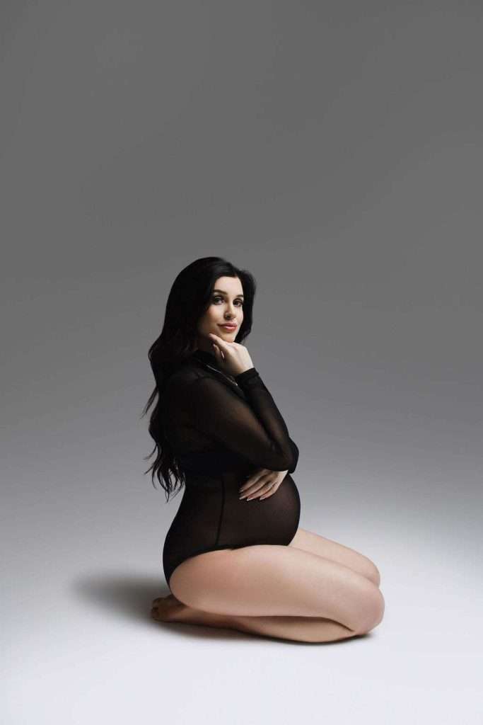 Pregnant woman in a sheer black bodysuit kneels on the floor, one hand on her belly and the other touching her chin, against a plain gray background.