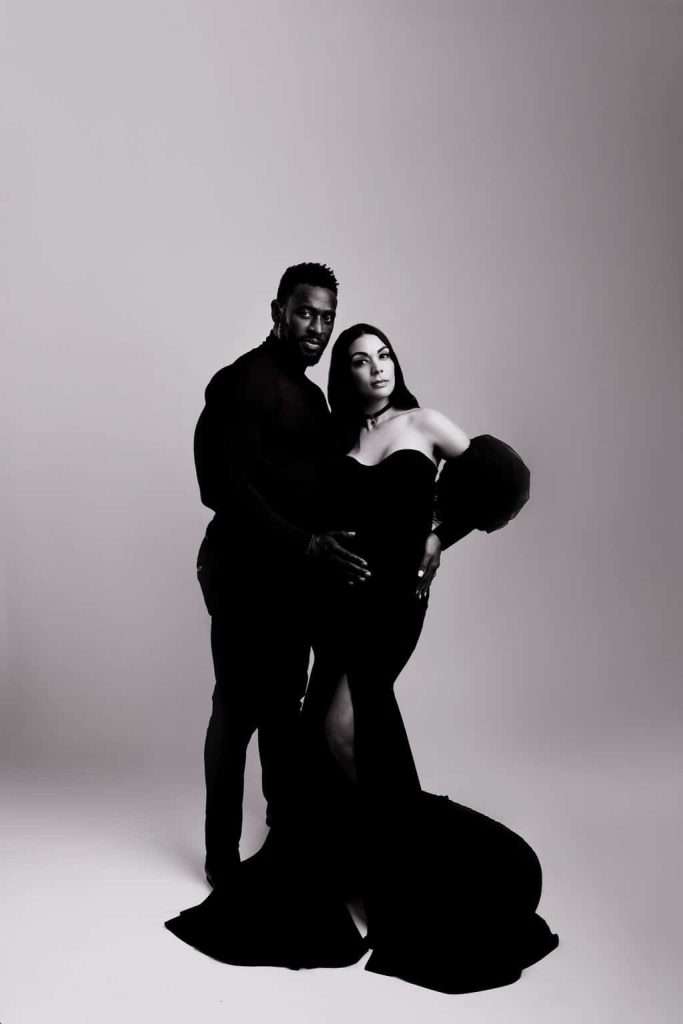 A man and a woman in formal black attire pose together in a studio setting; the woman is wearing a long gown and holding her stomach.