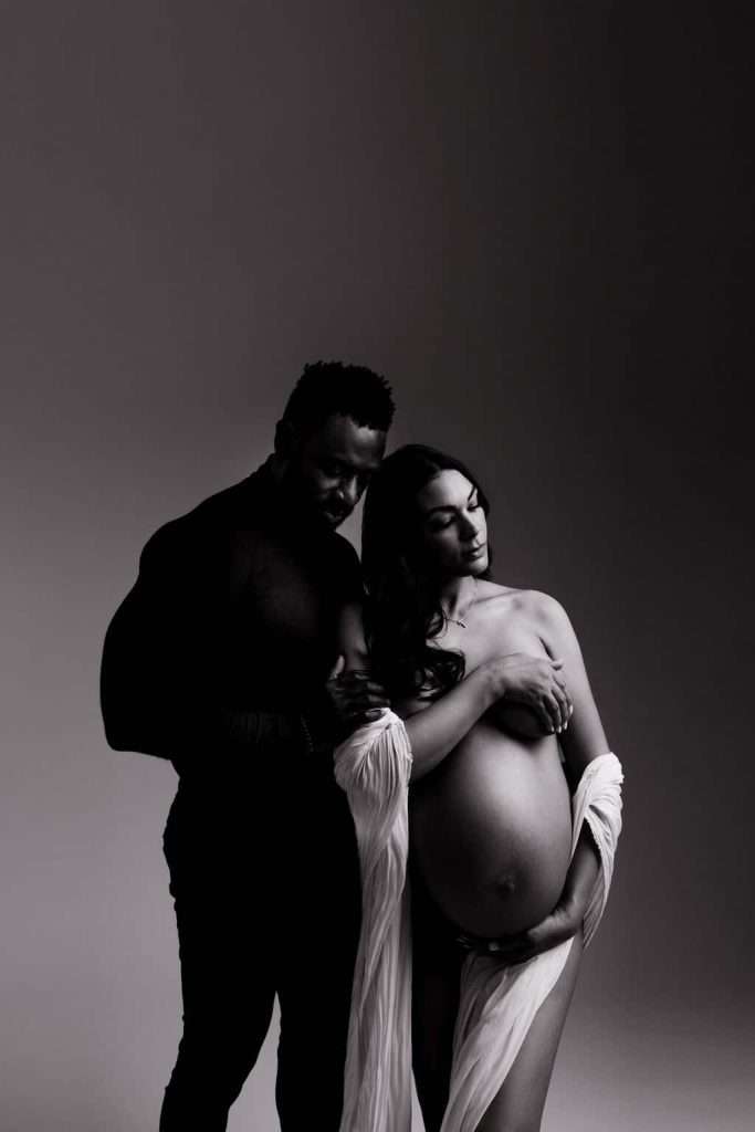 A man stands behind a pregnant woman draped in fabric, holding her arm and shoulder. The image is in black and white with dramatic lighting.