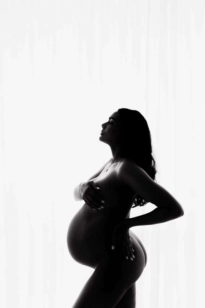 Silhouetted profile of a pregnant woman posing nude against a bright white background, one hand on her belly and the other covering her chest.