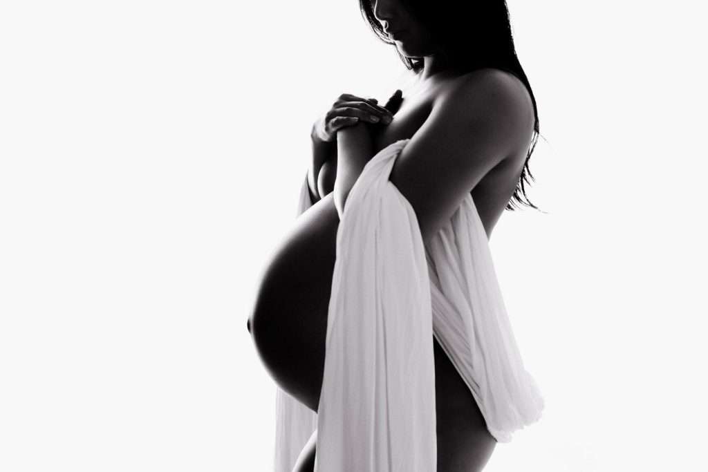 Silhouetted pregnant woman draped in a sheer fabric, holding it over her chest, with her baby bump visible against a white background.