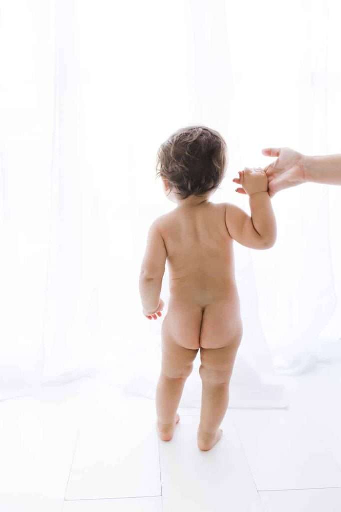A naked toddler stands facing a bright window with sheer curtains, holding an adult’s hand reaching in from the right.