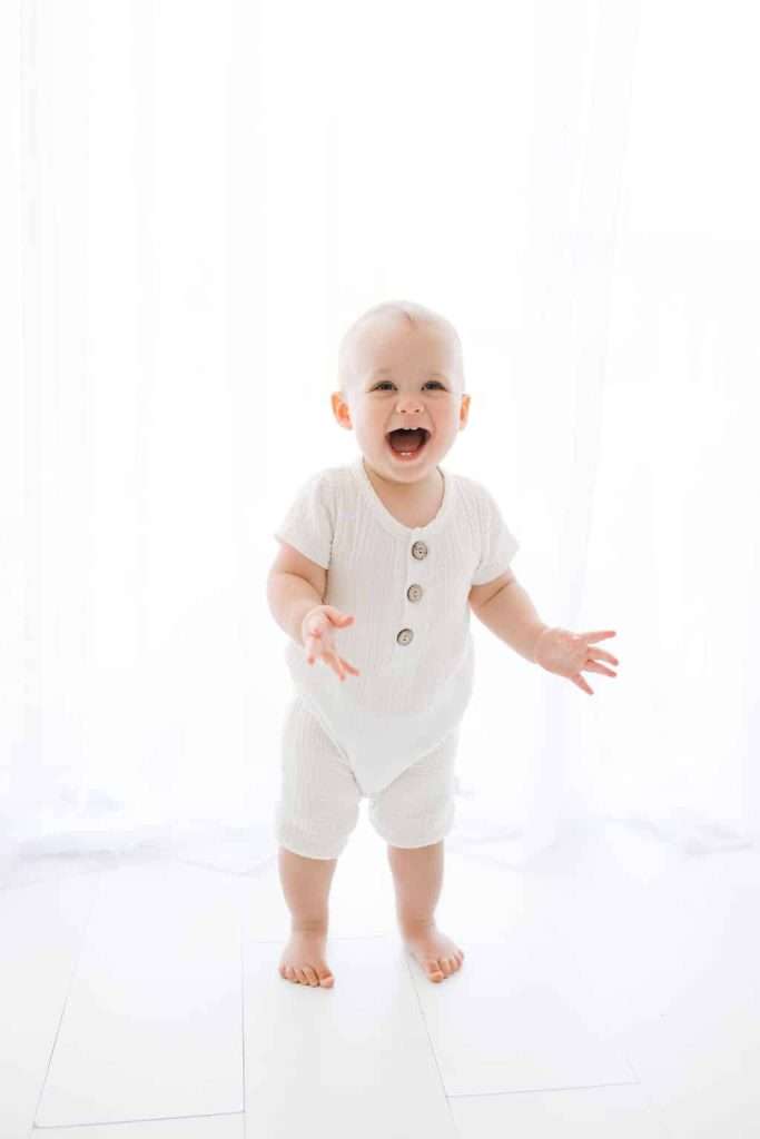 Smiling baby in a white outfit stands barefoot on a white floor with a bright, light-filled background.