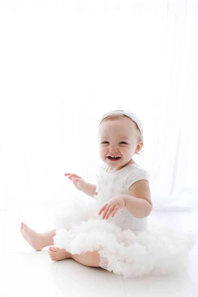 Smiling baby wearing a white dress and headband sits on a white floor in front of a bright, sheer curtain background.
