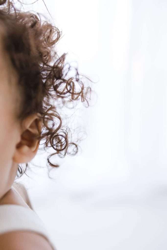 Close-up of a young child’s left ear and curly brown hair against a soft, light background.