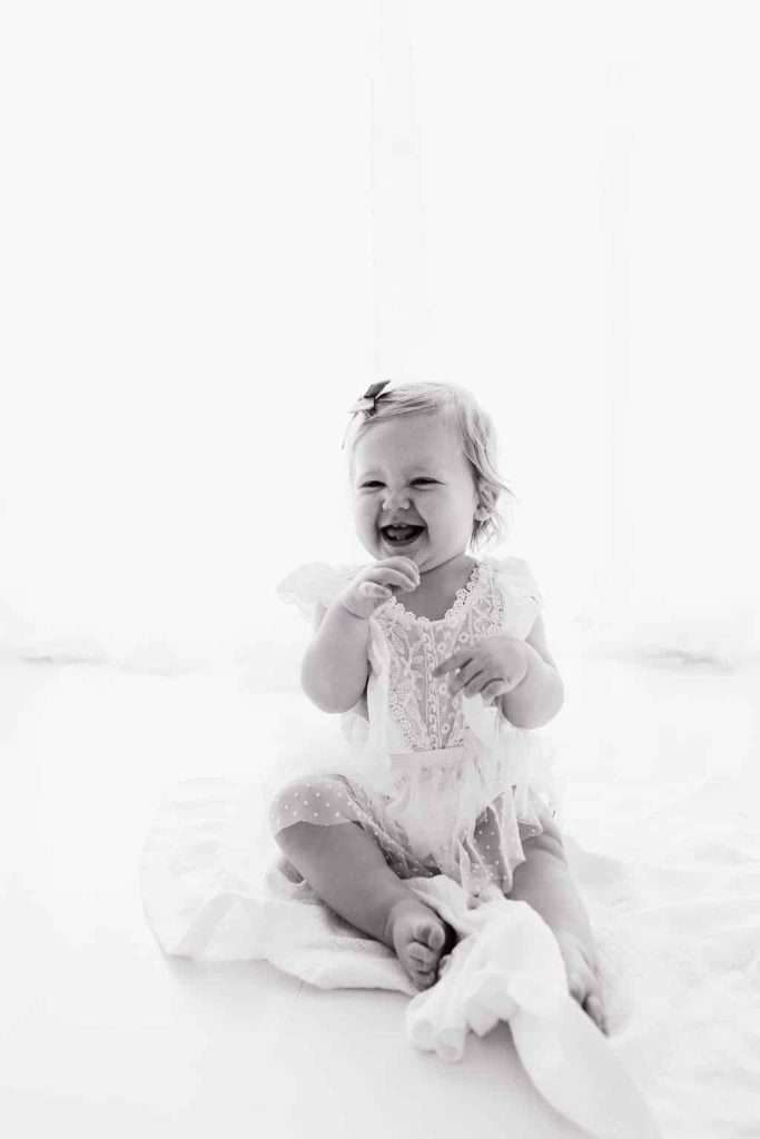 A baby in a lacy dress sits on a blanket, smiling and holding fabric, with a light background.