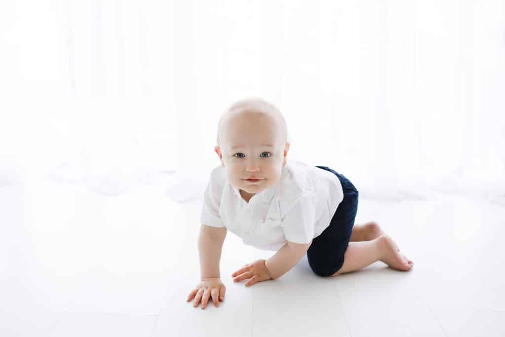 A baby wearing a white shirt and dark shorts is crawling on a white floor, looking forward. The background is bright and softly lit.