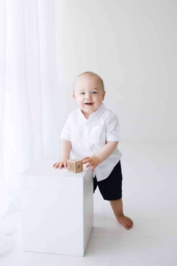 A baby in a white shirt and dark shorts stands by a white cube, holding a wooden block. The background is bright and minimal.