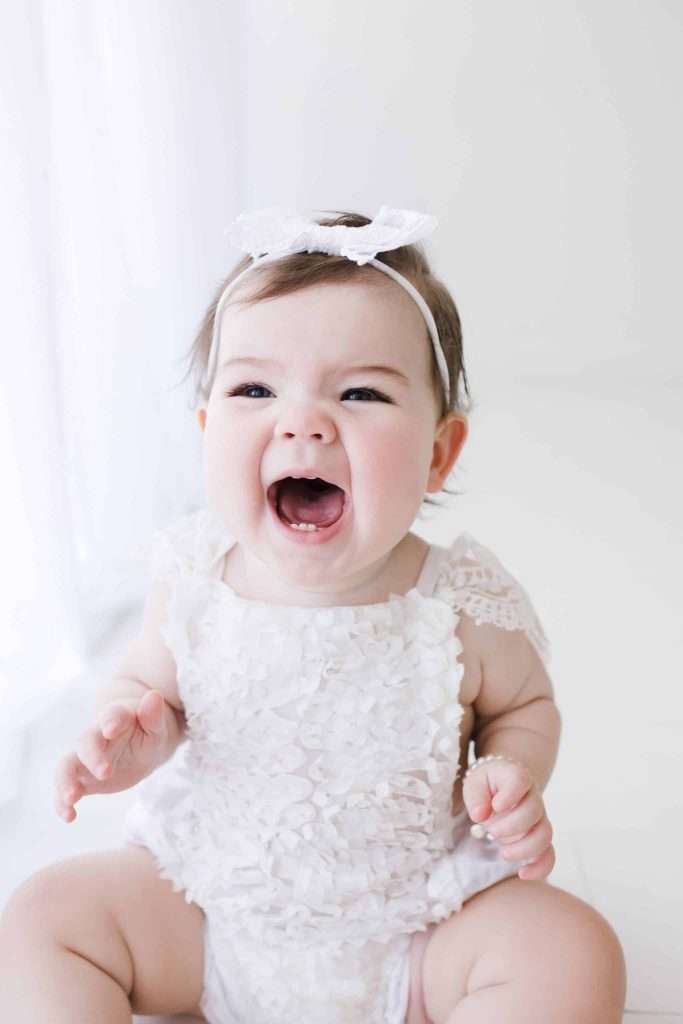 A baby wearing a white outfit and headband sits indoors, mouth open wide as if laughing or shouting.