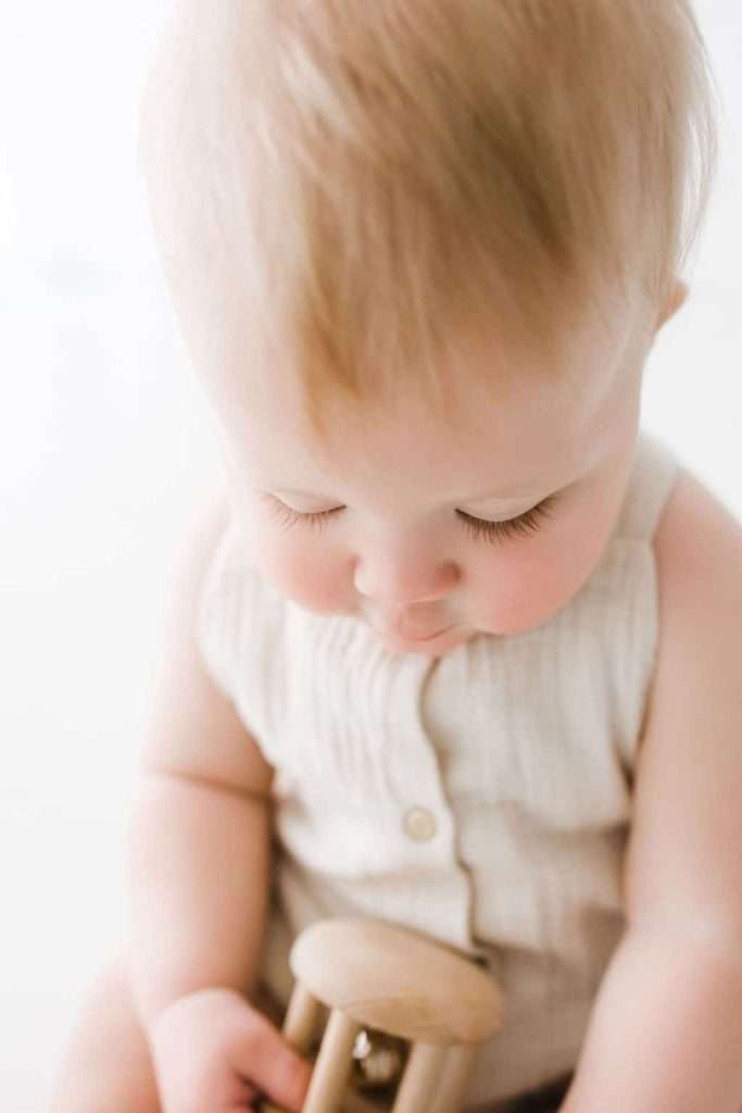 Baby with light hair looks down while holding a small wooden rattle, wearing a sleeveless light-colored outfit.