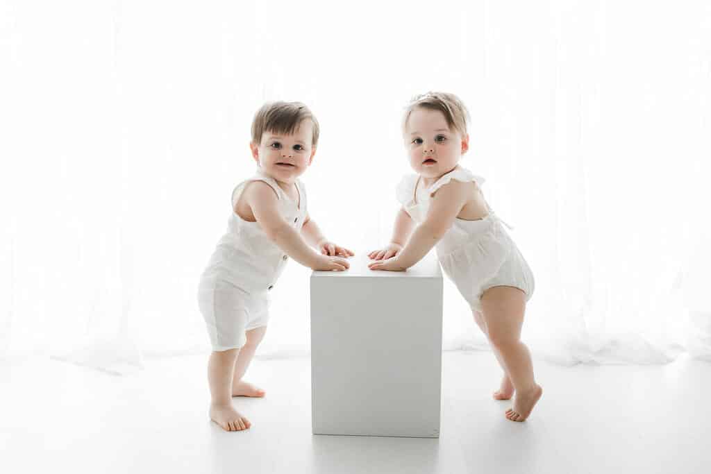 Two babies in white outfits stand barefoot on a white floor, each with one hand resting on a white cube. The background is bright and softly lit.