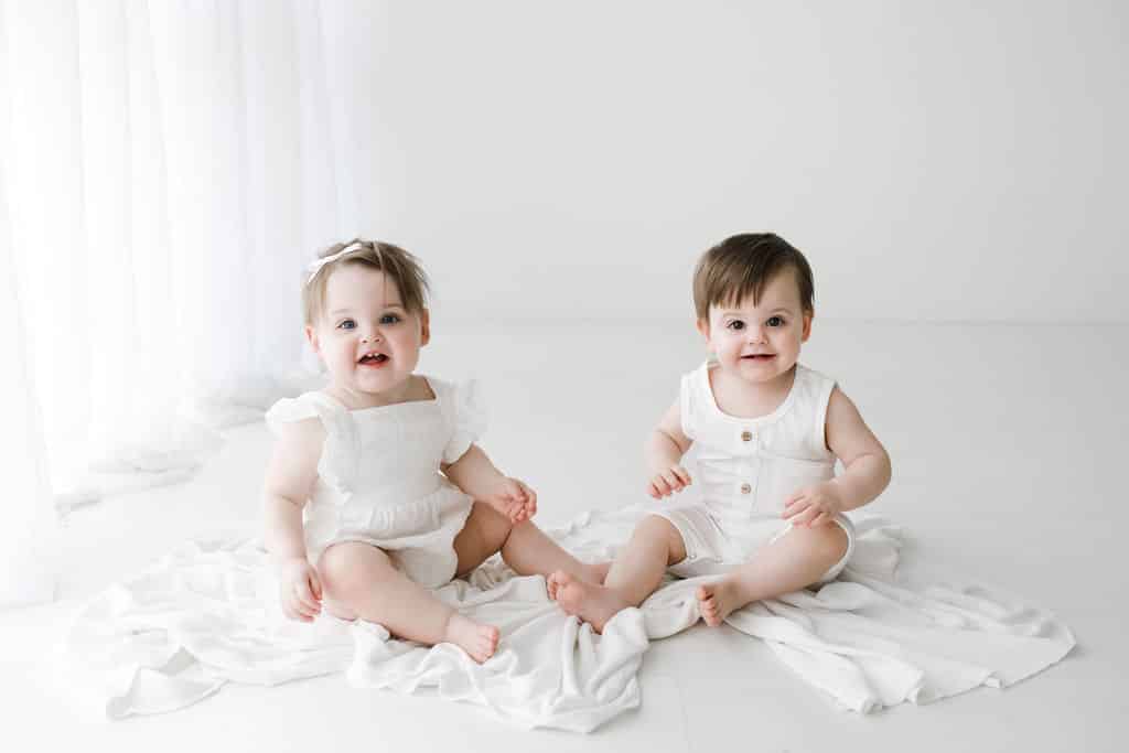 Two babies in white outfits sit on a white blanket in a bright, minimal room with light curtains and a white background.