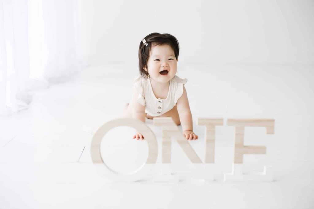 A baby in a white outfit crawls on a white floor, smiling, with large wooden letters spelling “ONE” placed in front of her.