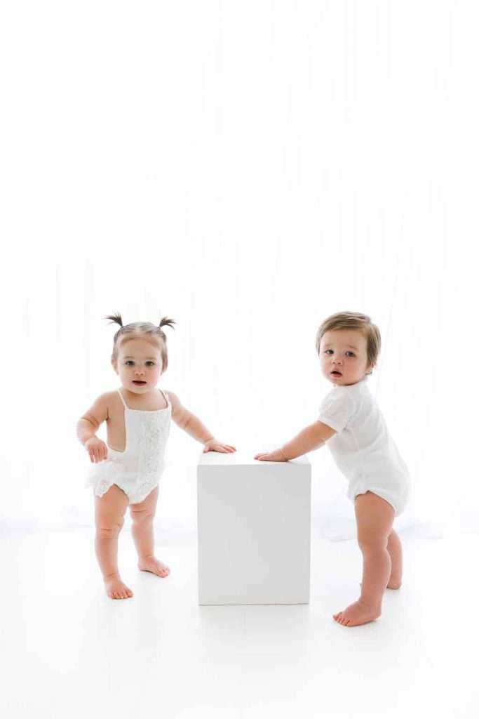 Two toddlers wearing white outfits stand next to a white cube in a brightly lit, minimalist setting with a white background.