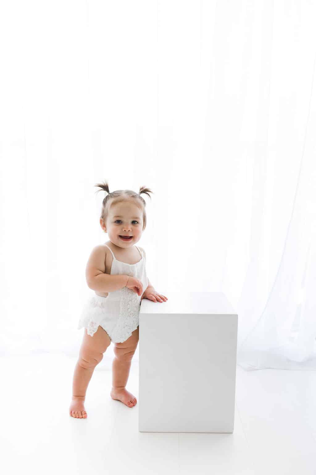 A toddler with pigtails, wearing a white outfit, stands next to a white cube against a bright white background.
