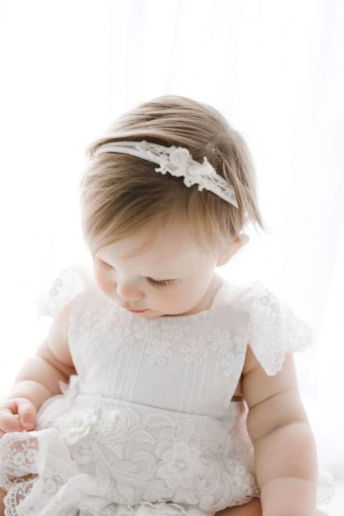 Baby wearing a white lace dress and floral headband looks down, sitting against a softly lit white background.