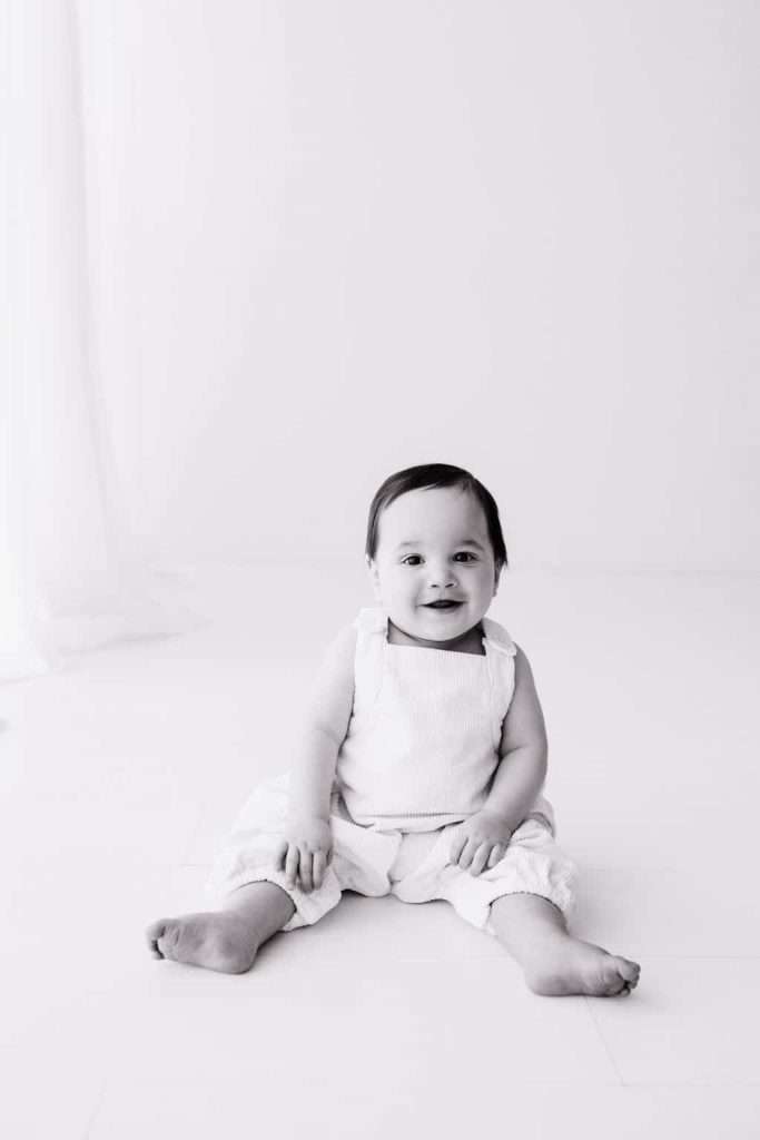 A baby sits on the floor in a light-colored room, wearing a sleeveless top and pants, looking at the camera and smiling.