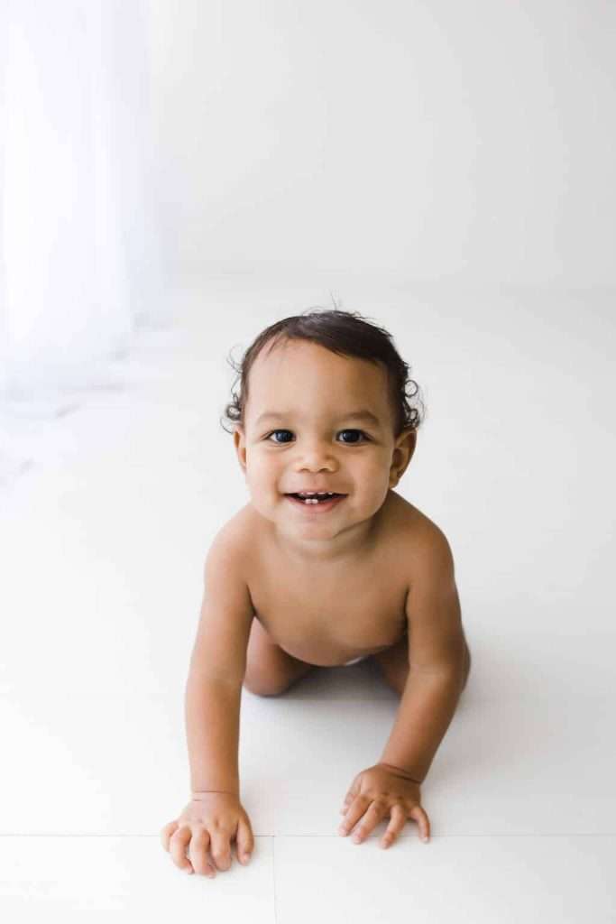 A baby with dark hair is crawling on a white floor, looking up and smiling at the camera in a brightly lit room.